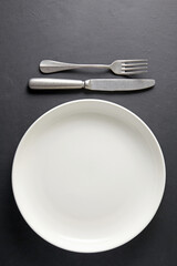 White ceramic plate and cutlery fork and knife on a black concrete background. Template for displaying food on menu