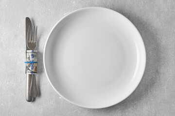 White ceramic plate on a gray concrete table and cutlery fork and knife wrapped in a hundred dollar bill. Top view with copy space