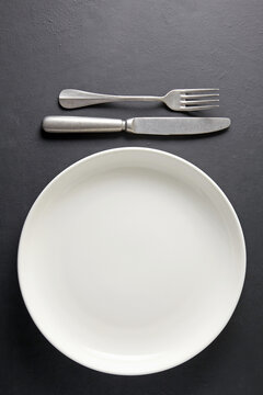 White ceramic plate and cutlery fork and knife on a black concrete background. Template for displaying food on menu
