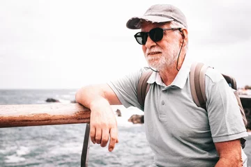 Fototapete Kanarische Inseln Portrait of senior traveler man with cap and sunglasses sitting at sea, elderly white haired bearded male carrying backpack enjoying summer vacation