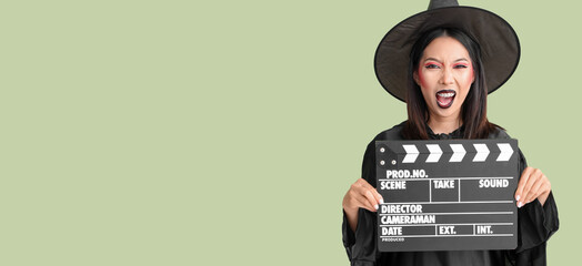 Asian woman dressed for Halloween as witch with clapperboard on green background with space for text