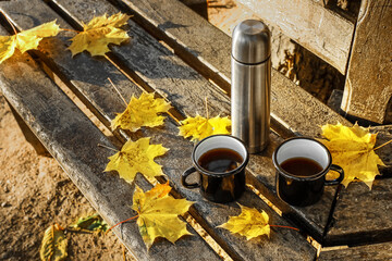 Fototapeta na wymiar Thermos with cups of coffee and fallen leaves on bench in autumn park