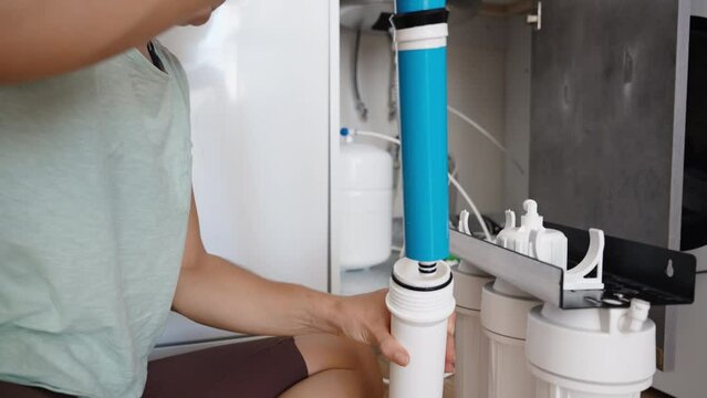 A young woman is independently changing water filters. She is pulling out a blue membrane filter from a white housing while sitting on the kitchen floor.