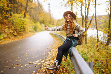Woman traveler in hiking clothes with a backpack and a hat stands on the side of the road, trying to catch a car on the road. Active weekend. Concept of adventure, travel, hitchhiking. © maxbelchenko