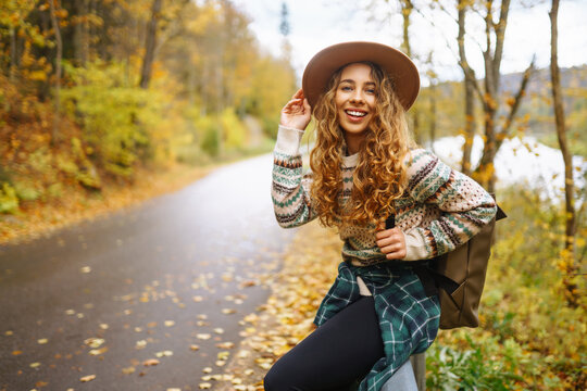 Woman traveler in hiking clothes with a backpack and a hat stands on the side of the road, trying to catch a car on the road. Active weekend. Concept of adventure, travel, hitchhiking.