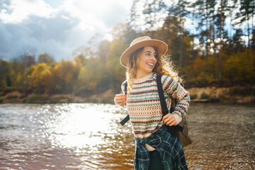 Female traveler in hiking boots with a backpack and a hat explores nature. A woman stands on the beach near the river enjoying the scenery and sunny weather, feeling freedom.