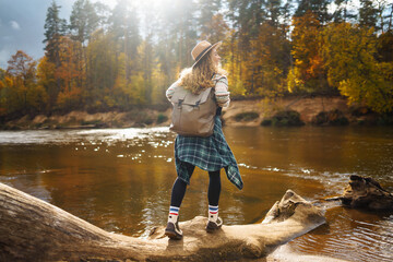 Female traveler in hiking boots with a backpack and a hat explores nature. A woman stands on the beach near the river enjoying the scenery and sunny weather, feeling freedom.