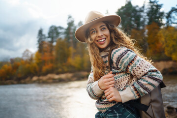 Female traveler in hiking boots with a backpack and a hat explores nature. A woman stands on the beach near the river enjoying the scenery and sunny weather, feeling freedom.