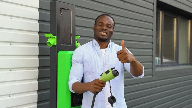 Portrait of smiling black man with a charger at EV charging station