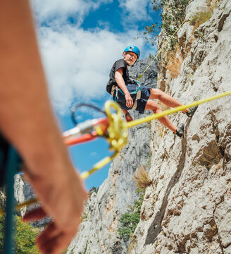 Teenager boy on the natural cliff climbing wall. Boy hanging on a rope using a climbing harness and the father belaying him on the ground using a belay device. Happy parenting and sports concept image