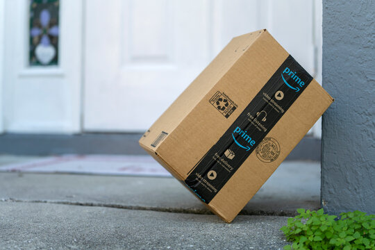 Box with Amazon logo in front of house door. Ordered online christmas gifts in cardboard package on doormat. Amazon Prime priority delivery. E-commerce concept. Sarasota, USA - December 19, 2022.