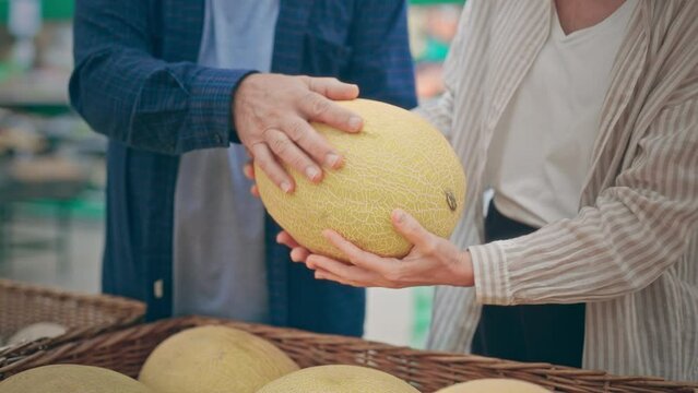 A man and a woman choose a ripe melon in a supermarket. Close-up of only hands. Unrecognizable