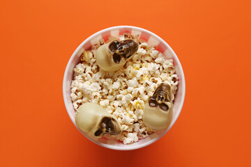 Bucket with tasty popcorn and skulls for Halloween celebration on orange background