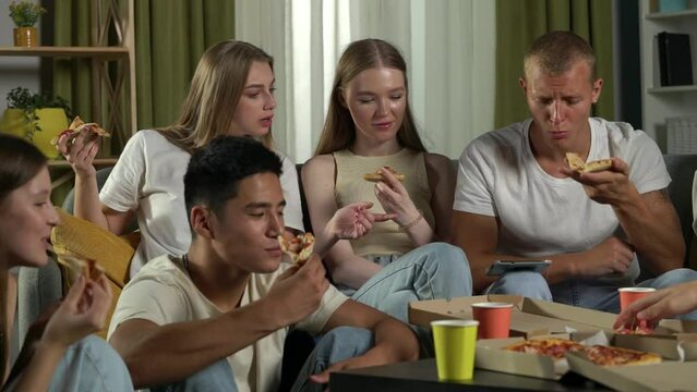 Medium video of a group of teens, young people, friends sitting on a couch and floor around the table, eating pizza, drinking beverages, chatting, talking, joking around.