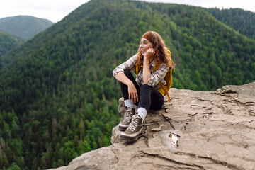 Beautiful traveler with a yellow hiking backpack observes the mountain scenery from a cliff, a view...