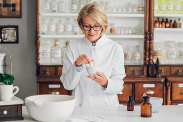 A mature pharmacist is standing in a vintage drugstore and mixing a cure in a mortar