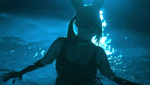 Sensual woman in leather BDSM mask moving seductively in swimming pool water under neon color light. Halloween party, attractive chick masquerade bunny, nighttime games for adults