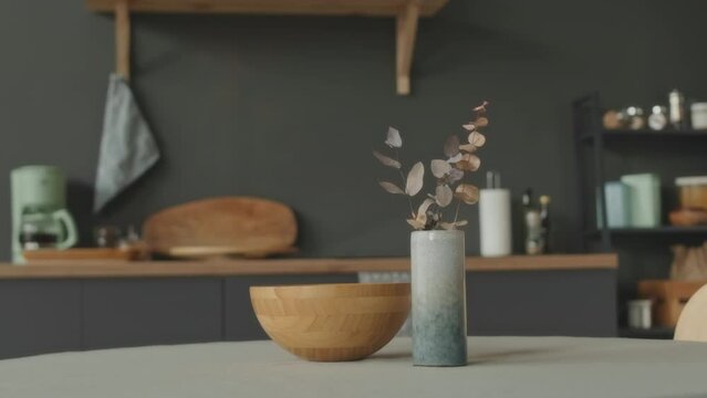 No people shot of dry flower arrangement in epoxy vase and wooden bowl on white kitchen table in minimalist apartment with graphite grey walls
