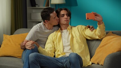 Shot of two homosexual men, couple at home. They are sitting on the couch, hugging, taking selfies, expressing love and affection to each other.