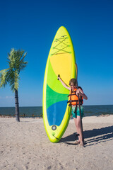 happy 10 year old boy holding a green sup board and paddle for sup boarding on a sunny day at the seaside.