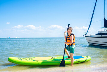 a little boy in an orange life jacket is about to ride his green SUP board in the sea. A 10-year-old child stands with one foot on the board, the other in the sea, holding an oar. on summer vacation w