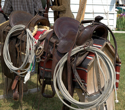 Two western saddles on wooden saddle stands, with lariats, and saddle blankets in place, set for a cowboy or other rider western style.
