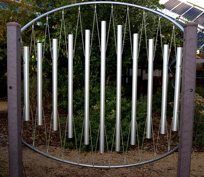 Acoustical cylinders for creating musical tones hang from an arched support in an outdoor setting.
