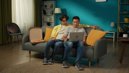 Full-size shot of a homosexual couple at home. They are sitting on the couch, watching something on a laptop, eating pizza, hugging and talking warmly to each other.