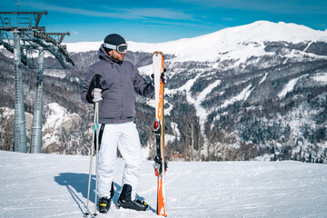 Young male skier standing on top of mountain while holding skies. Athletic man in full winter sport...