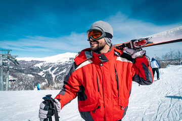 Portrait of young male smiling skier standing on top of mountain while holding skies. Joyful man posing on ski slope covered with snow during the winter and looking away. - obrazy, fototapety, plakaty