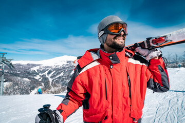 Portrait of confident male skier in red jacket standing on top of mountain while holding skies....