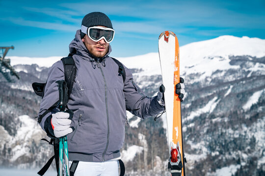 Portrait of young man skier standing on top of mountain while holding skies and poles. Man wearing warm winter clothes hiking and skiing high in the mountains.