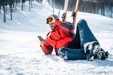 Smiling male skier resting on snow taking a break while using phone. Satisfied confident man wearing red winter jacked and taking ski goggles off lying down on a ski slope and texting on cell phone.