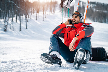 Smiling male skier sitting on snow taking a break while using smart phone and laughing. Handsome...