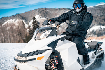 Fototapeta na wymiar Man driving a snowmobile. Male person in full winter equipment sitting on snowmobile and looking at camera in the mountains covered with bright white snow.