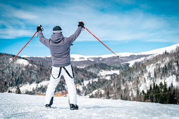 Young professional skier on mountain top ready to ride down - winner stance - hands raised up. Male...