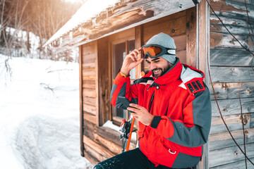 Young smiling male skier using smart phone in front of log cabin. Fashionable man in full ski equipment checking his mobile phone on vacation. - obrazy, fototapety, plakaty