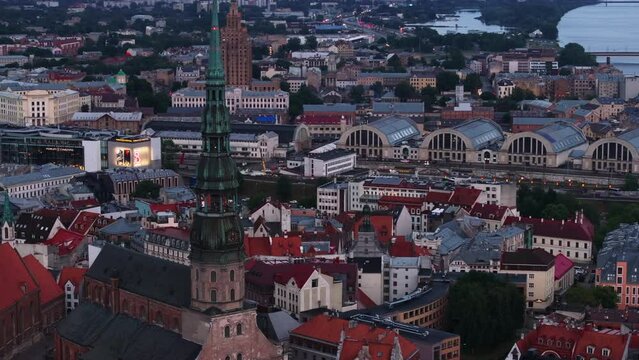 Aerial cinematic footage of historic city centre with tall church tower. Pavilions of large Riga Central Market and other tourist sights at dusk. Riga, Latvia