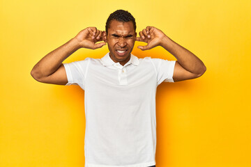 Fototapeta na wymiar African American man in white polo, yellow studio, covering ears with fingers, stressed and desperate by a loudly ambient.