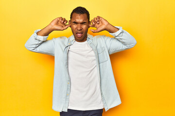 Fototapeta na wymiar African American man in blue shirt, yellow studio, covering ears with fingers, stressed and desperate by a loudly ambient.