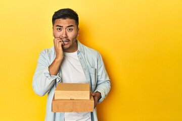 Asian man holding delivery food boxes, yellow studio backdrop biting fingernails, nervous and very...