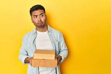 Asian man holding delivery food boxes, yellow studio backdrop confused, feels doubtful and unsure.