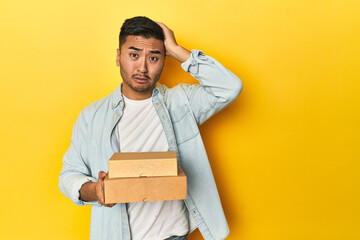 Asian man holding delivery food boxes, yellow studio backdrop being shocked, she has remembered important meeting.