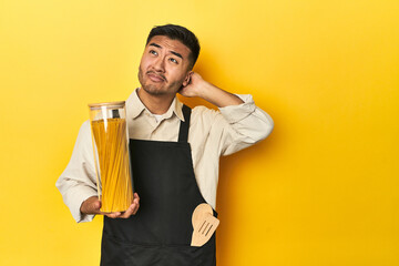 Asian chef with spaghetti jar, yellow studio backdrop touching back of head, thinking and making a...