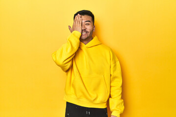 Bold Asian man in yellow hoodie on yellow studio having fun covering half of face with palm.