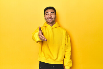 Bold Asian man in yellow hoodie on yellow studio stretching hand at camera in greeting gesture.