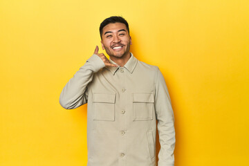 Fototapeta premium Asian man in stylish gray shirt on yellow studio showing a mobile phone call gesture with fingers.