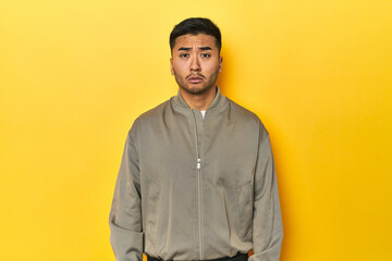 Stylish Asian man in gray jacket on yellow studio sad, serious face, feeling miserable and...