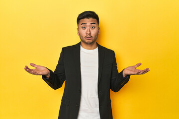 Asian man in black business jacket on yellow studio doubting and shrugging shoulders in questioning...
