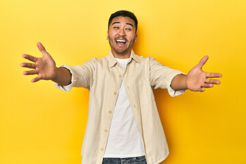 Casual Asian man with open shirt, white tee on yellow studio feels confident giving a hug to the camera.